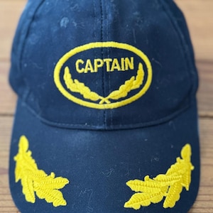 May include: Navy blue baseball cap with the word "CAPTAIN" in yellow, inside a yellow oval with yellow leaf accents. Yellow leaf accents are also on the brim of the cap. The cap is made of a durable fabric.