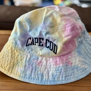 May include: A tie-dye bucket hat with the text "CAPE COD" embroidered in black. The hat features a mix of pastel colors, including blue, pink, yellow, and white. The hat is made of a soft fabric and has a wide brim.