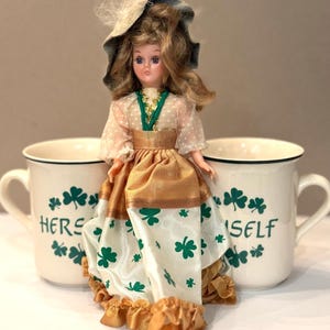 May include: A vintage doll with a hat and a gold and white dress, adorned with green accents, sits between two white mugs. The mugs feature green shamrocks and the words "HERE" and "SELF".