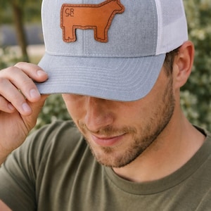 May include: Gray and white trucker hat with a brown leather patch featuring a cow silhouette. The hat has a gray front panel, a white mesh back, and a curved brim. The patch has the letters "CR" on the cow.