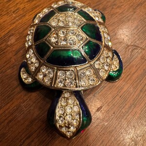 May include: A jeweled turtle brooch with a gold-tone metal frame. The turtle's shell is a mosaic of green and blue enamel, accented with clear rhinestones. The brooch has a classic, elegant design.