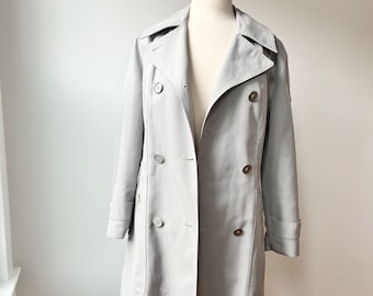 May include: A light gray trench coat with a double breasted closure and a belt. The coat has a classic design and is made of a lightweight fabric.