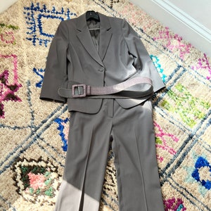 May include: A gray suit with a matching blazer and pants. The suit has a single button closure and a belt with a large buckle.