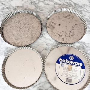 Puede incluir: Cuatro moldes redondos para tartas con bordes estriados. Los moldes son de metal plateado. Uno de los moldes tiene una etiqueta que dice "bakeware" y "strawberry meringue tart". Los moldes están sobre una superficie de mármol.