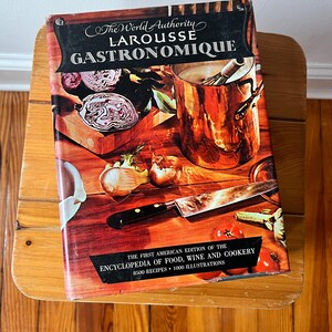 May include: A hardcover cookbook titled "Larousse Gastronomique" rests on a wooden stool. The cover features a vibrant illustration of cooking ingredients and tools. Text reads "The World Authority" and "Encyclopedia of Food, Wine and Cookery".