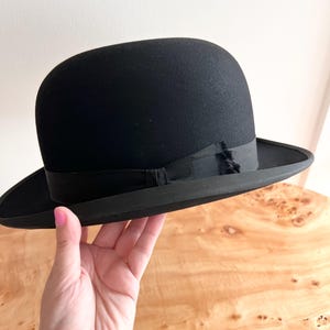 Könnte beinhalten: Ein schwarzer Bowlerhut mit einer abgerundeten Krone und einem schmalen Rand. Der Hut hat ein schwarzes Band am unteren Rand der Krone. Dieser klassische Hut wird oft mit formeller Kleidung in Verbindung gebracht. Der Hut hat einen Durchmesser von ca. 14 cm.