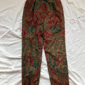 May include: Paisley print trousers in shades of red, green, and brown. The pants feature a high waist and a classic, tailored fit. The intricate paisley pattern covers the entire surface of the trousers, creating a vintage aesthetic. The trousers are laid flat on a white surface.