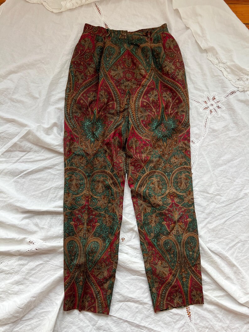 May include: Paisley print trousers in shades of red, green, and brown. The pants feature a high waist and a classic, tailored fit. The intricate paisley pattern covers the entire surface of the trousers, creating a vintage aesthetic. The trousers are laid flat on a white surface.