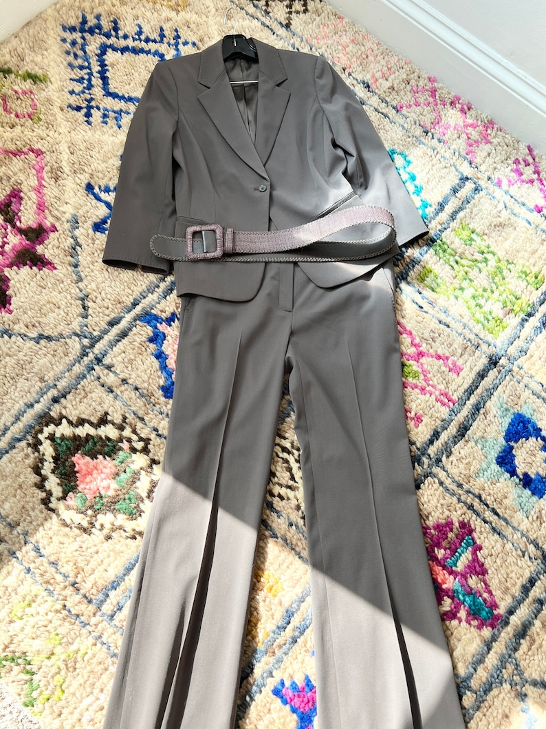 May include: A gray suit with a matching blazer and pants. The suit has a single button closure and a belt with a large buckle.