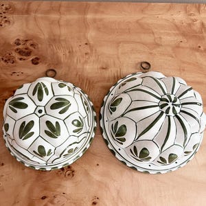 May include: Two white ceramic molds with green leaf designs. Each mold has a rounded shape with a scalloped edge and a metal loop for hanging. The molds are displayed on a wooden surface.