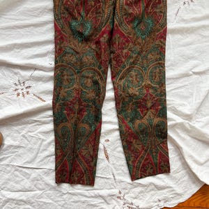 May include: Paisley print pants in shades of red, green, and brown. The pants feature a detailed, symmetrical pattern. The fabric appears to be lightweight, with a slight sheen. The pants are laid flat on a white surface.