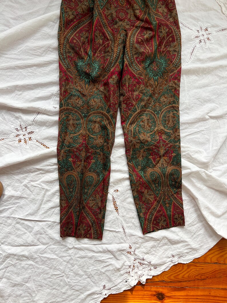 May include: Paisley print pants in shades of red, green, and brown. The pants feature a detailed, symmetrical pattern. The fabric appears to be lightweight, with a slight sheen. The pants are laid flat on a white surface.