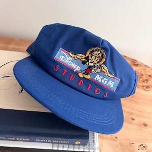 May include: A royal blue baseball cap featuring an embroidered patch with the text Disney MGM Studios and an illustration of Mickey Mouse holding a film clapperboard, with a lion emblem above the text on a light blue rectangular background.
