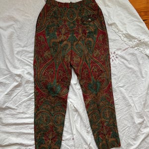 May include: High-waisted pants with a vibrant paisley pattern in red, green, and gold. The pants feature a buttoned pocket and a tailored fit, showcasing a vintage aesthetic. The fabric appears to be a lightweight material.