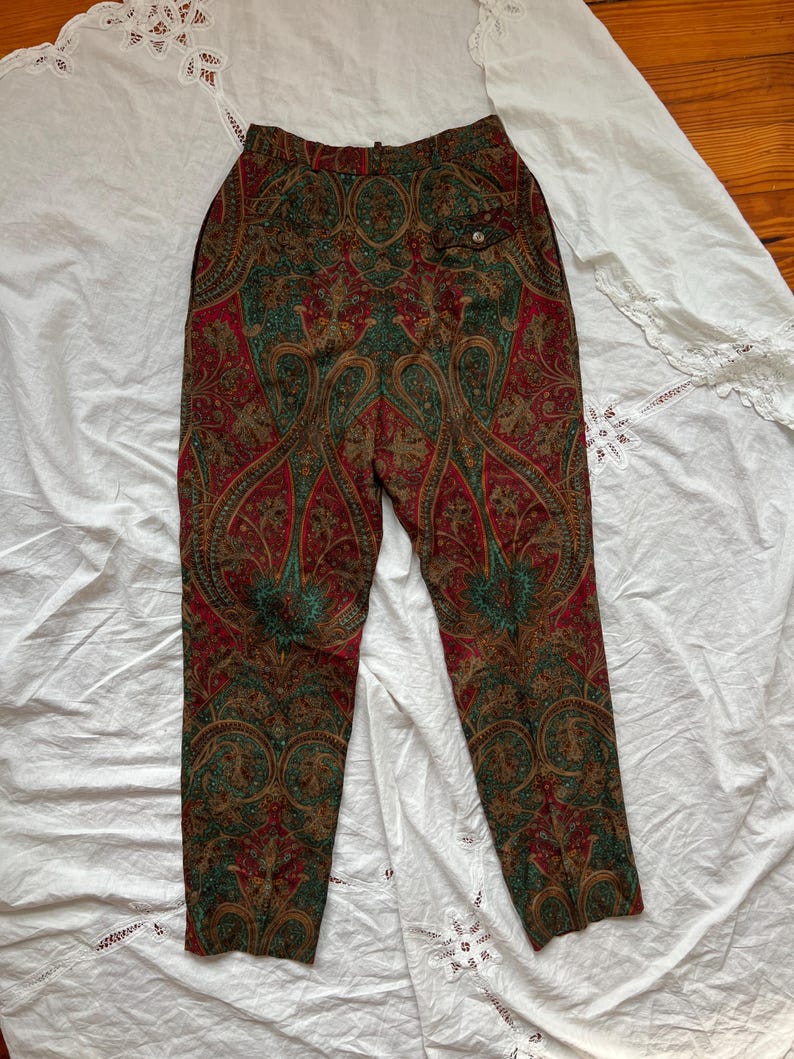 May include: High-waisted pants with a vibrant paisley pattern in red, green, and gold. The pants feature a buttoned pocket and a tailored fit, showcasing a vintage aesthetic. The fabric appears to be a lightweight material.