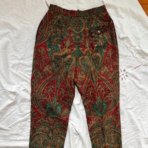 May include: Paisley print pants in red, green, and gold. The pants feature a high waist and a button closure. The intricate paisley pattern covers the entire surface of the pants, creating a vintage aesthetic. The pants are made of a lightweight fabric.