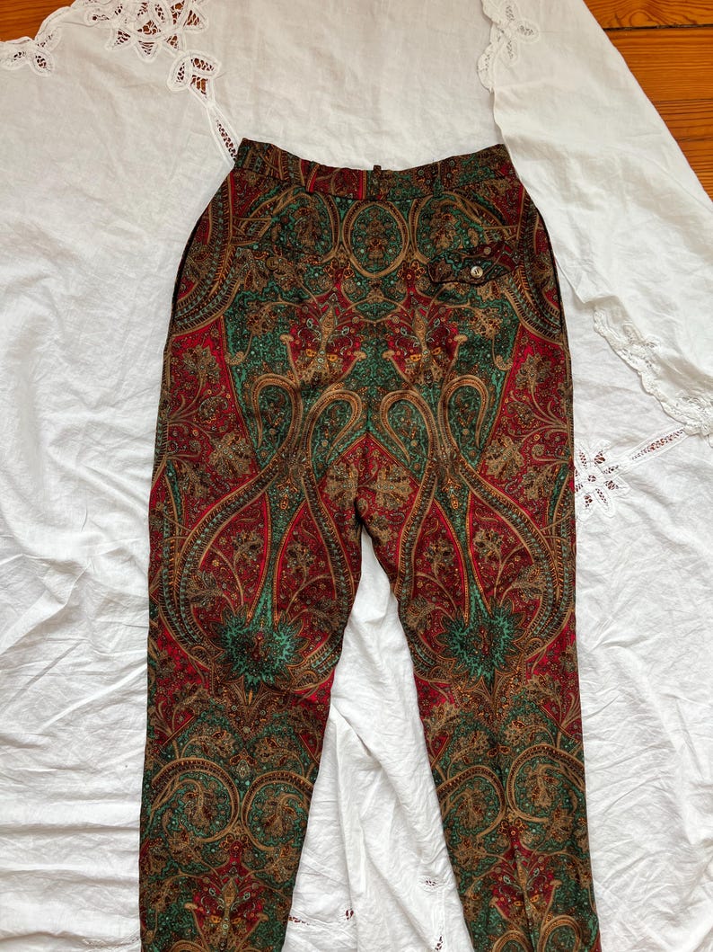 May include: Paisley print pants in red, green, and gold. The pants feature a high waist and a button closure. The intricate paisley pattern covers the entire surface of the pants, creating a vintage aesthetic. The pants are made of a lightweight fabric.