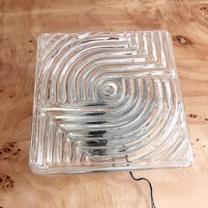 May include: A square glass light fixture featuring a clear, textured surface with concentric, swirling patterns radiating from a central point. The glass panel has a thick, molded design with a black electrical cord extending from the bottom edge.