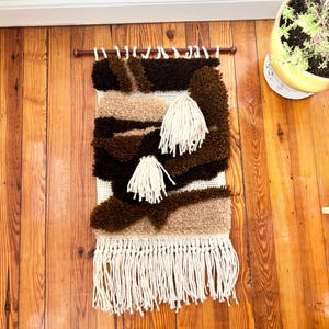 May include: A woven wall hanging with a brown, tan, and cream color scheme. The textile features a textured design with a fringe at the bottom and two cream-colored tassels. The wall hanging is mounted on a wooden rod.
