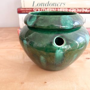 Op de afbeelding: Een glanzende, groene keramische pot met een afgeronde vorm en een uitlopende rand. De pot heeft een gat aan de zijkant en staat op een houten oppervlak. Boeken zijn op de achtergrond gestapeld. De pot is waarschijnlijk voor decoratief of functioneel gebruik.