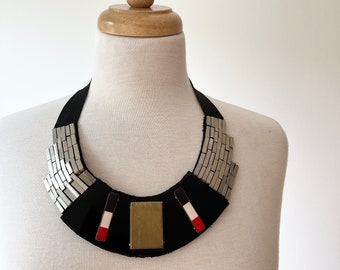 Puede incluir: Un collar llamativo con una base negra y detalles metálicos plateados. El collar presenta un rectángulo central de color latón y detalles rojos, blancos y negros. Se exhibe en un maniquí blanco.