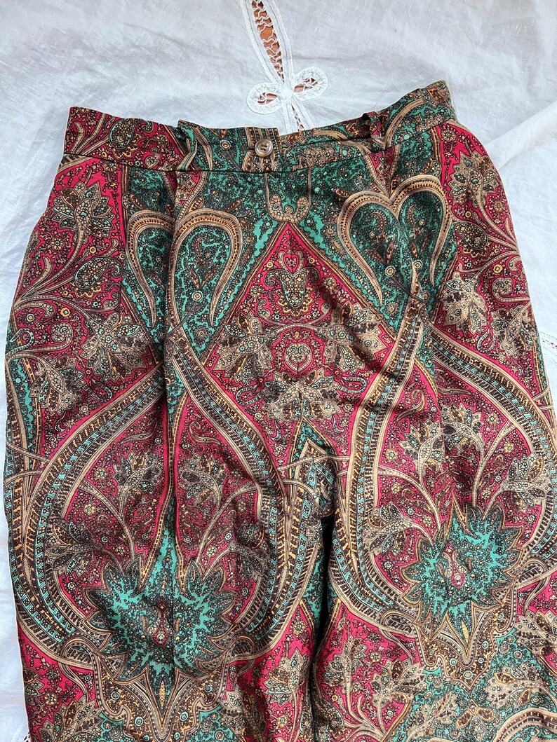 May include: High-waisted pants with a vibrant paisley pattern in shades of red, teal, and brown. The fabric features intricate, detailed designs. The pants have a button and zipper closure.