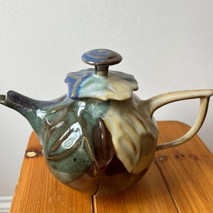 Pode incluir: Uma chaleira de cerâmica artesanal com um design floral. A chaleira tem um corpo arredondado com uma alça e um bico. A tampa tem um pequeno botão. As cores incluem tons de verde, castanho e bege, com detalhes em azul. A chaleira está numa superfície de madeira.