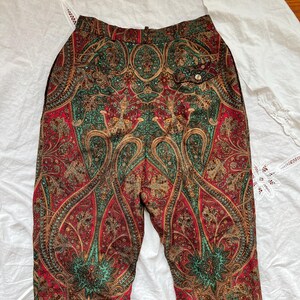 May include: Paisley print pants in shades of red, green, and gold. The pants feature a high waist and a button closure. The intricate pattern is visible across the entire garment, creating a vintage aesthetic. The pants are made of a soft, flowing fabric.