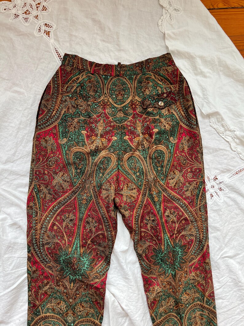 May include: Paisley print pants in shades of red, green, and gold. The pants feature a high waist and a button closure. The intricate pattern is visible across the entire garment, creating a vintage aesthetic. The pants are made of a soft, flowing fabric.