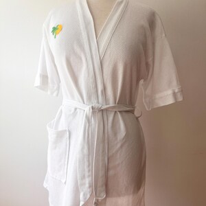 May include: A white waffle-knit robe featuring short sleeves, a tie-waist belt, and a single patch pocket. A small, embroidered yellow and green palm tree graphic is positioned on the upper left chest area of the garment.