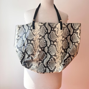 May include: A large tote bag featuring a grey, black, and white snakeskin pattern. The bag has two black shoulder straps attached with gold-toned metal hardware. The overall shape is wide and slouchy, displaying a textured reptilian print.