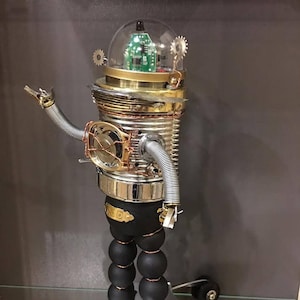 Può includere: Scultura di robot fatta a mano costruita con metallo riciclato e componenti elettronici. Il robot presenta una testa a cupola trasparente con un circuito stampato, un corpo cilindrico e braccia articolate. Le gambe sono fatte di sfere nere e rotola su una base con ruote.