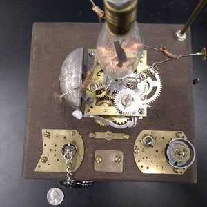 May include: A steampunk-style clockwork sculpture with a light bulb, gears, and a bell on a wooden base.