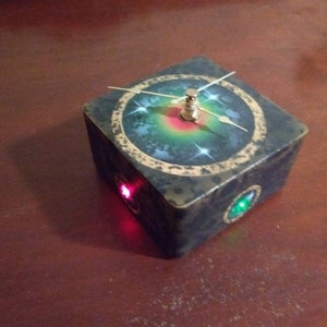 May include: A black wooden cube clock with a gold-rimmed clock face featuring a green and red galaxy design. The clock has a gold hand and a gold knob. There are two small lights on the cube, one red and one green.