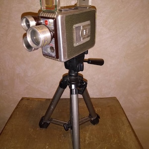 May include: A vintage silver and black 8mm film camera mounted on a black tripod. The camera has a viewfinder and a lens with a red ring. The camera is on a wooden surface.