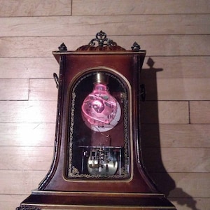 Puede incluir: Un reloj de madera antiguo con un frente de vidrio. La esfera del reloj presenta un orbe de vidrio rosa y giratorio. El reloj está sobre una superficie de madera.