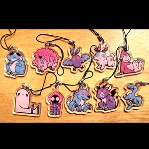 May include: A collection of nine clear acrylic charms featuring colorful cartoon illustrations of various creatures, including a blue frog, a pink blob, a moth, a rabbit, a gorilla, a ghost, a hooded figure, a wolf, a demon, and a sea serpent.