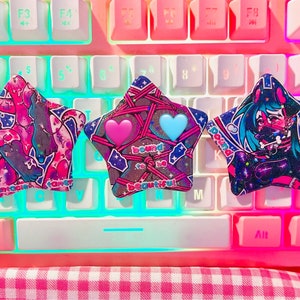 Könnte beinhalten: Drei sternförmige Tastenkappen mit einzigartigen Designs liegen auf einer rosa-weißen Tastatur. Die Tastenkappen zeigen farbenfrohe Illustrationen, darunter eine Figur, Herzen und den Text "bound to be beautiful". Die Tastatur hat weiße und rosa Tasten.