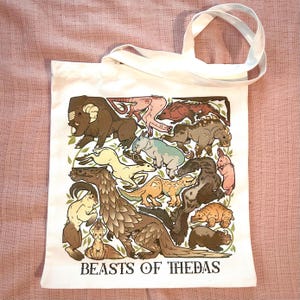 Könnte beinhalten: Weißer Stoffbeutel mit einer farbenfrohen Illustration verschiedener Fantasietiere. Das Design umfasst einen Widder, ein Nashorn und andere Tiere, mit dem Text "BEASTS OF THEDAS" unten. Die Tasche hat weiße Griffe.