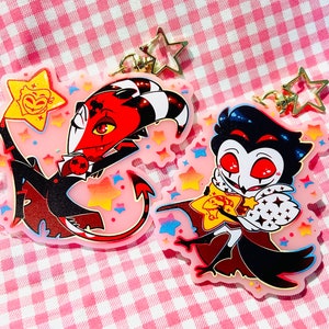 May include: Two cartoon character keychains. One keychain features a red and black demon with a star. The other keychain features a dark blue and white character with a star. Both keychains have star-shaped clasps and are on a pink background.