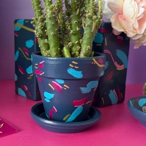 May include: A dark blue terracotta plant pot with a matching saucer, featuring hand-painted abstract brushstrokes in bright pink, light blue, and gold. A green cactus with small spines is planted inside the pot, which has a matte finish.