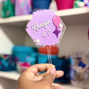 May include: A purple and clear acrylic cake topper with the text "Khadija is 6". The topper has a hexagon shape with a balloon and star design. The topper is held by a clear stick.