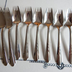 May include: A collection of eight silver-colored forks arranged on a white surface with a decorative border. The forks have a classic design with a three-tine head and a slender, tapered handle. The forks are made of a shiny, reflective metal.