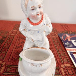 May include: A vintage porcelain figurine of a child kneeling, wearing a white dress with blue star accents and a red beaded necklace. The figurine has a small bowl and is on a white base.