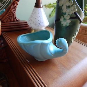 May include: A light blue ceramic cornucopia-shaped planter with a smooth, glossy finish. The planter is sitting on a wooden surface.