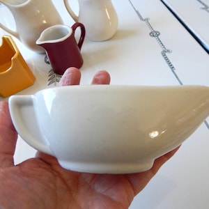 May include: A white ceramic gravy boat with a handle is held in a hand. Other ceramic serving pieces in cream, burgundy, and orange are in the background. The gravy boat has a spout and a curved handle.