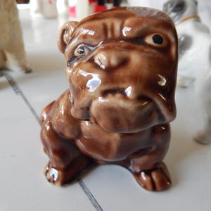 May include: A brown ceramic bulldog figurine with a glossy finish. The bulldog is in a seated position, with a detailed face and visible wrinkles. Other dog figurines are visible in the background.