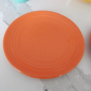 May include: A bright orange ceramic dinner plate with a slightly raised rim and concentric circles in the center.