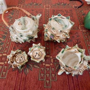 May include: A ceramic tea set with a seashell design. The set includes a teapot with a woven handle, a sugar bowl, a creamer, and two salt and pepper shakers. The pieces are cream-colored with green and brown accents, and sit on a red patterned tablecloth.