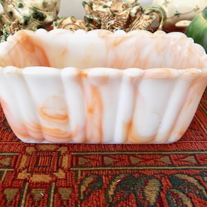 May include: A rectangular, white and orange swirled glass planter with a scalloped rim. The planter is on a patterned red surface, with other decorative items in the background.